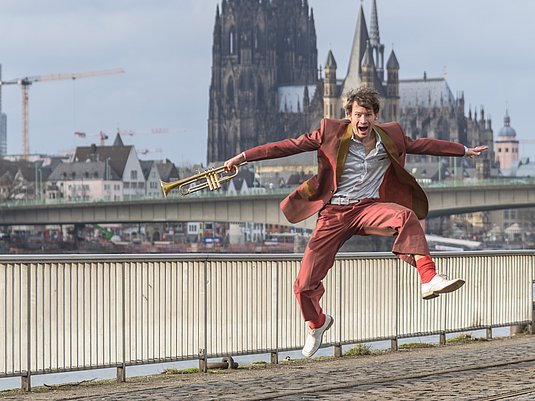 Matthias Schriefl Person mit einer Trompete in der Hand springt in die Luft. Dahinter ist der Kölner Dom zu sehen