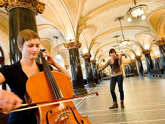 Zwei Studierene spielen Violoncello und Violine in einem historischen Gebäude.
