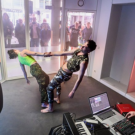 Zwei Tänzer*innen und ein DJ treten in einem verschlossen Raum vor Publikum auf