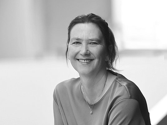 Das Bild zeigt eine freundlich lächelnde Frau mit rotbraunen Haaren, die sich an ein weißes Geländer lehnt.