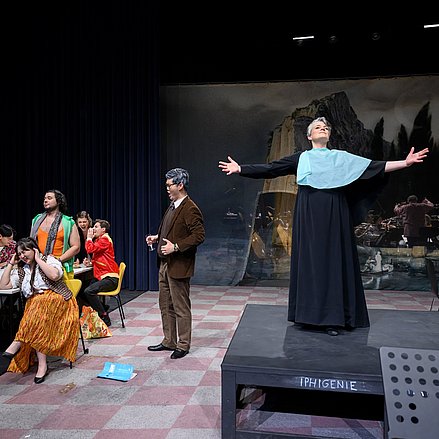 Das Bild zeigt eine Theaterszene mit mehreren Schauspielerinnen und Schauspielern auf einer Bühne. 