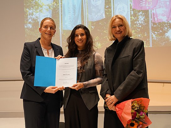 Drei Personen auf der Bühne mit einer Urkunde und einem Blumenstrauß