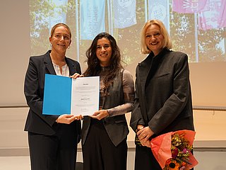 Drei Personen auf der Bühne mit einer Urkunde und einem Blumenstrauß