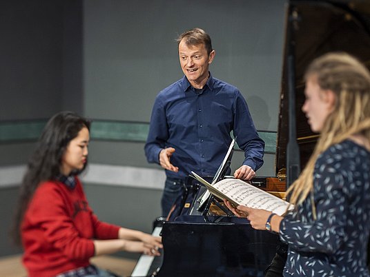 Professor mit zwei Studierenden beim Gesangsunterricht mit einem Klavier.