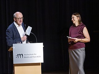 Zwei Personen hinter einem Rednerpult. Die eine Person hält einen Sparten in der Hand. 