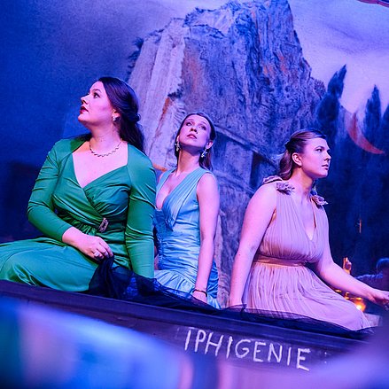Drei Darstellerinnen in eleganten Kleidern sitzen auf einer Bühne mit der Aufschrift „IPHIGENIE“ 