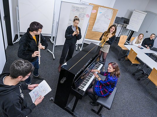 Mehrere Studierende stehen mit Instrumenten um ein Klavier mit einer Professorin