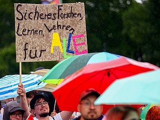 Person hält in der CSD-Parade ein Schild hoch mit dem Text: Sicheres Forschen, Lernen, Lehren für Alle