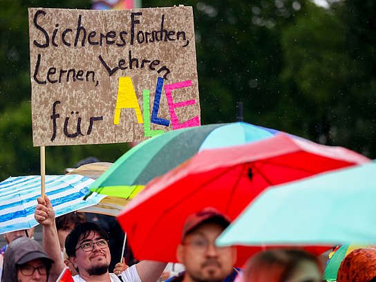 Person hält in der CSD-Parade ein Schild hoch mit dem Text: Sicheres Forschen, Lernen, Lehren für Alle