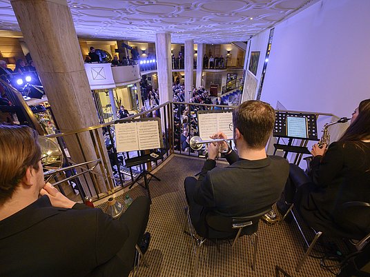 Drei Studierende sitzen auf einer Empore und spielen Trompete und Posaune vor Publikum.