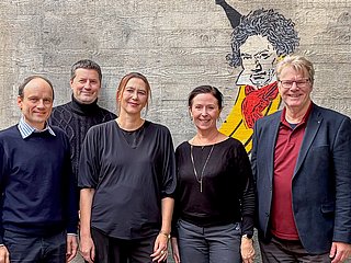 Vier Personen (Kanzler*in, Prorektor*in und Rektor*in) des Rektorats stehen vor einer Betonwand.