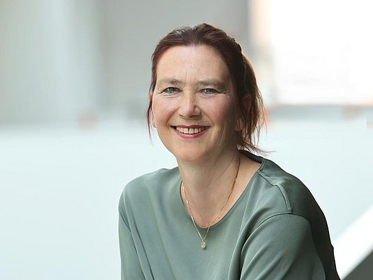 Das Bild zeigt eine freundlich lächelnde Frau mit rotbraunen Haaren, die sich an ein weißes Geländer lehnt.