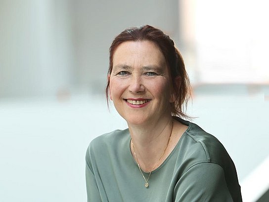 Das Bild zeigt eine freundlich lächelnde Frau mit rotbraunen Haaren, die sich an ein weißes Geländer lehnt.