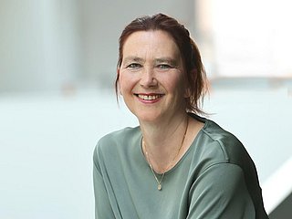 Das Bild zeigt eine freundlich lächelnde Frau mit rotbraunen Haaren, die sich an ein weißes Geländer lehnt.