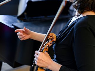 Person steht vor einem Klavier und hält eine Violine in der Hand.