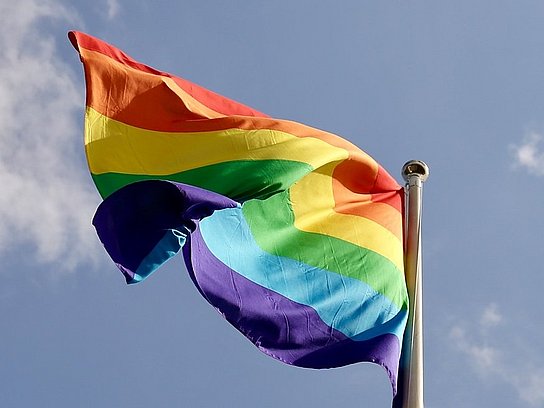 Ein Flaggenmast mit einer Regenbogenfahne und blauem Himmel