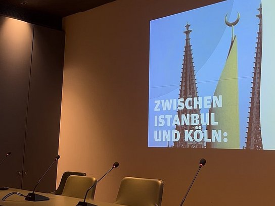 Tisch mit drei Stühlen, Mikrofone auf dem Tisch und an der Wand eine Präsentation "Zwishcne Istanbul und Köln"