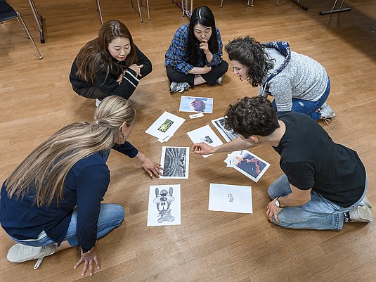 Gruppe Studierende mit Professorin sitzen auf dem Boden und schauen sich Ausdrucke an.