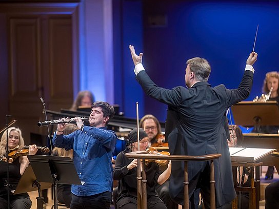 Eine Person spielt konzentriert Oboe auf einer Konzertbühne vor einem Orchester mit Dirigent