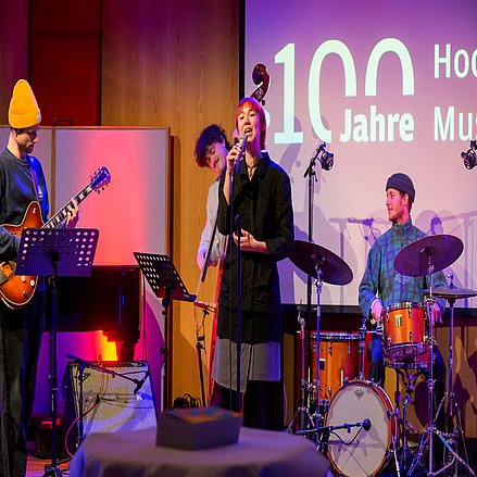 Vier Personen stehen auf der Bühne mit Mikrofon und Instrumenten