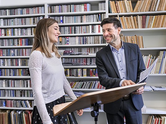 Zwei Personen stehen vor einem Notenständer und im Hintergrund ist ein Bücherregal