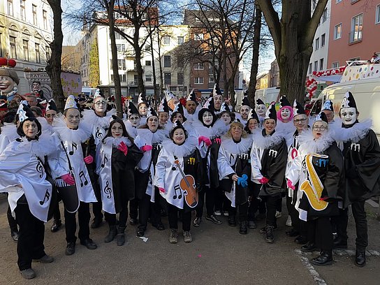Das Foto zeigt eine Gruppe von Menschen, die sich für einen Karnevalsumzug verkleidet haben. Sie tragen fantasievolle Pierrot-ähnliche Kostüme in Schwarz und Weiß, geschmückt mit Musiknoten und Instrumentenmotiven.