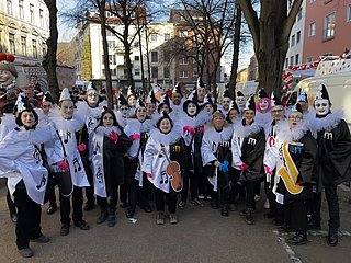 Das Foto zeigt eine Gruppe von Menschen, die sich für einen Karnevalsumzug verkleidet haben. Sie tragen fantasievolle Pierrot-ähnliche Kostüme in Schwarz und Weiß, geschmückt mit Musiknoten und Instrumentenmotiven.