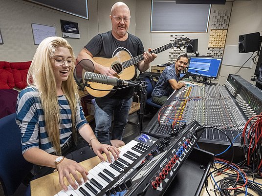 Unterrichtssituation Studierende am Keyboard mit Professor mit einer Gitarre im Tonstudio.