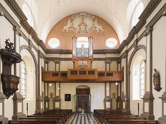 Es ist der Kirchenraum der Ursulinenkirchen zu sehen. Die Ahrend-Orgel ist auf der Empore positioniert, während unten die Kirchenbänke und mittig der Gang zu sehen ist.
