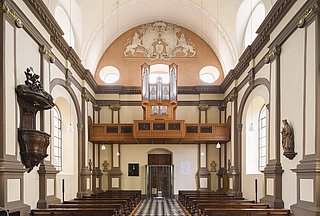 Es ist der Kirchenraum der Ursulinenkirchen zu sehen. Die Ahrend-Orgel ist auf der Empore positioniert, während unten die Kirchenbänke und mittig der Gang zu sehen ist.