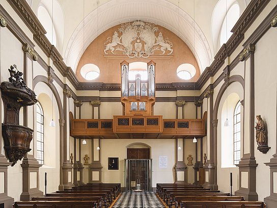 Orgel Ursulinenkirche Es ist der Kirchenraum der Ursulinenkirchen zu sehen. Die Ahrend-Orgel ist auf der Empore positioniert, während unten die Kirchenbänke und mittig der Gang zu sehen ist.