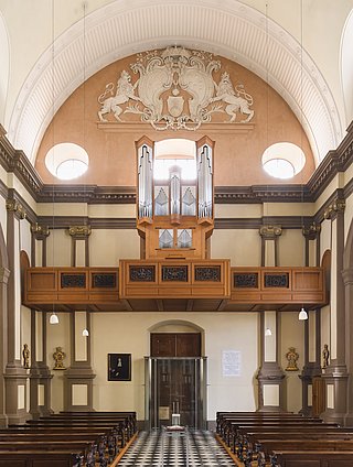 Es ist der Kirchenraum der Ursulinenkirchen zu sehen. Die Ahrend-Orgel ist auf der Empore positioniert, während unten die Kirchenbänke und mittig der Gang zu sehen ist.